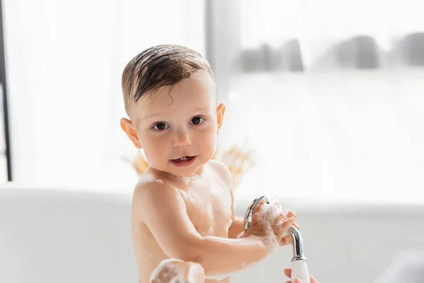 抱着淋浴头看着相机 身体上有泡泡浴泡沫的湿幼儿 — 图库照片