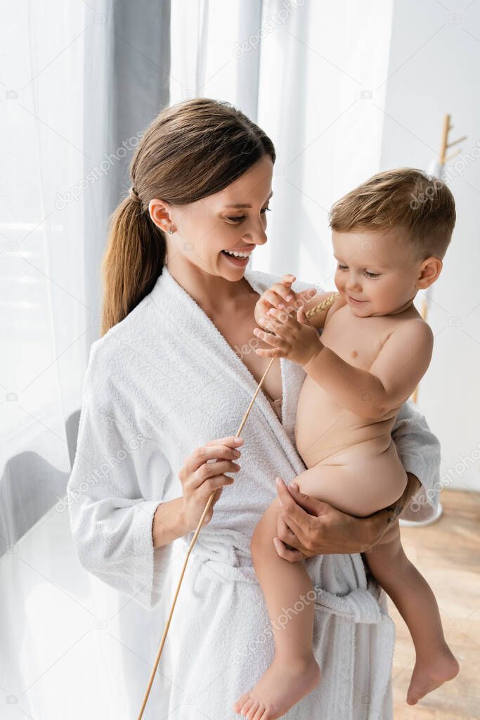 smiling mother in bathrobe holding wheat spikelet near nude toddler son 