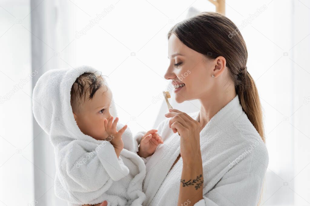 smiling mother brushing teeth and holding in arms toddler son in bathrobe 