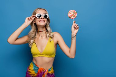 Sarı bikinili ve güneş gözlüklü gülümseyen genç kadın mavi üzerinde tatlı bir lolipop tutuyor.