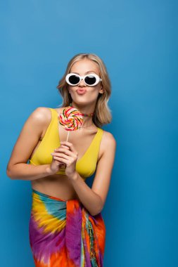 Sarı bikinili, güneş gözlüklü, dudaklarını büzen, mavi renkli lolipop tutan genç bir kadın.