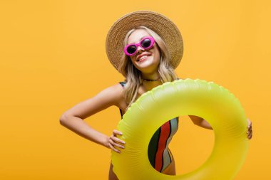 Güneş şapkalı ve mayo giymiş gülümseyen kadın sarıda izole edilmiş şişme yüzük tutuyor.