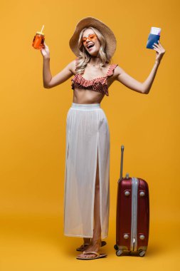 full length of happy woman in sun hat standing near luggage while holding passport and cocktail on yellow  clipart
