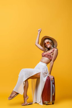 full length of happy woman in sun hat sitting on luggage and rejoicing on yellow  clipart