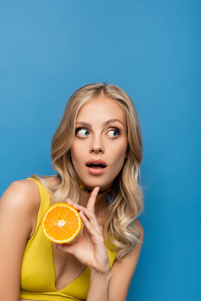 surprised young woman in yellow bikini top holding orange half isolated on blue