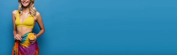 обрезанный вид веселой молодой женщины в желтом топе бикини и саронг держа оранжевую половину на голубом, баннер