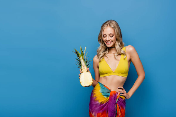 веселая молодая женщина в желтом бикини и саронг, держащая сладкий ананас наполовину на голубом