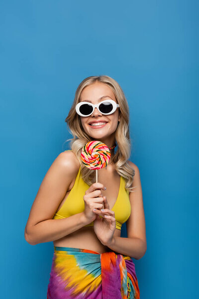 улыбающаяся молодая женщина в желтом топе бикини и солнечных очках, держащих сладкий леденец, изолированный на голубом