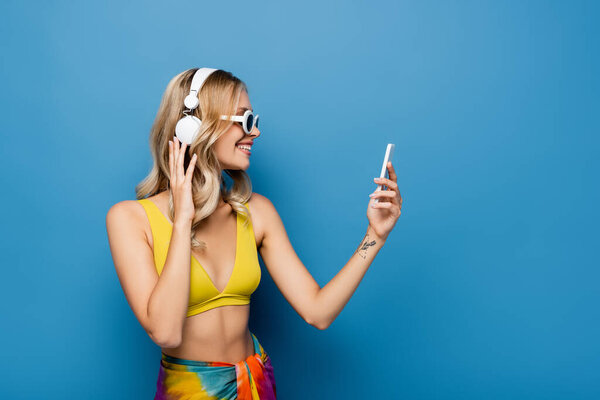 вид сбоку счастливой женщины в беспроводных наушниках и топе бикини с помощью смартфона синего цвета