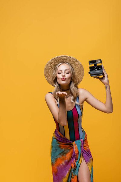 young woman in straw hat holding vintage camera and sending air kiss isolated on yellow