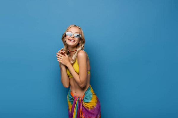 радостная молодая женщина в солнечных очках и бисером красочное ожерелье улыбаясь изолированы на синий