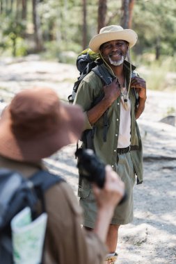 Sırt çantalı neşeli Afrikalı Amerikalı turist ormanda bulanık karısına bakıyor. 