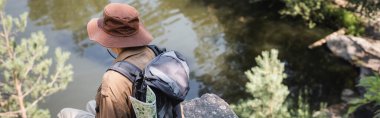 Sırt çantalı yaşlı turist göl kenarındaki kayalıklarda oturuyor. 