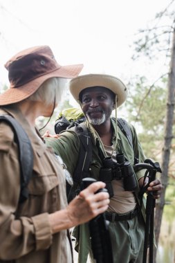 Neşeli Afro-Amerikan turist yürüyüş direkleri ve dürbünleriyle bulanık karısının yanında duruyor. 