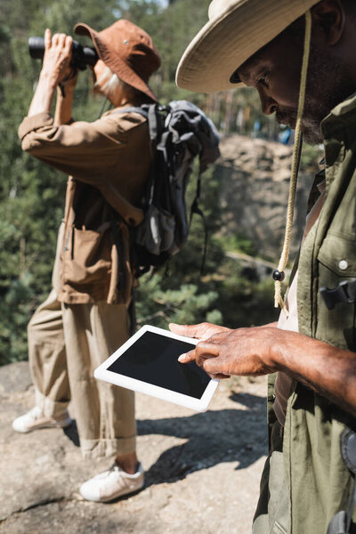 African american man using digital tablet near blurred wife with binoculars 