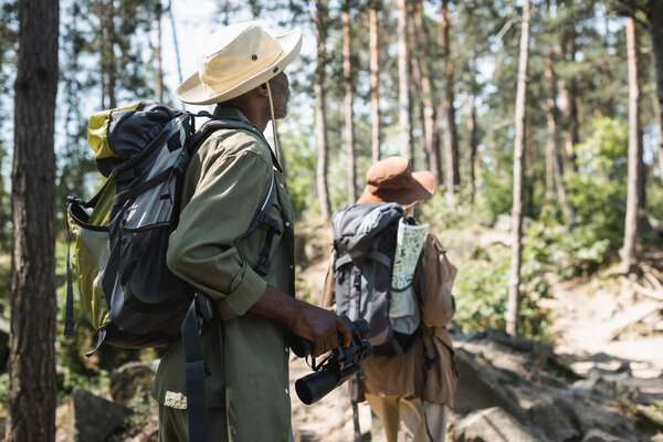 Африканский американский турист с рюкзаком и биноклем, идущий рядом с размытой женой в лесу 