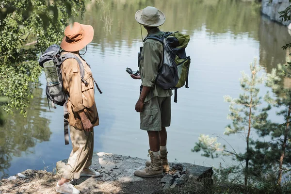 年龄较大的种族间夫妇 背着背包和指南针站在湖边 — 图库照片