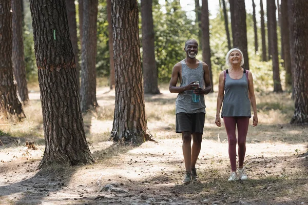 アフリカ系アメリカ人の夫の近くを歩く笑顔のシニアスポーツ女性スポーツボトルとともに森の中 — ストック写真