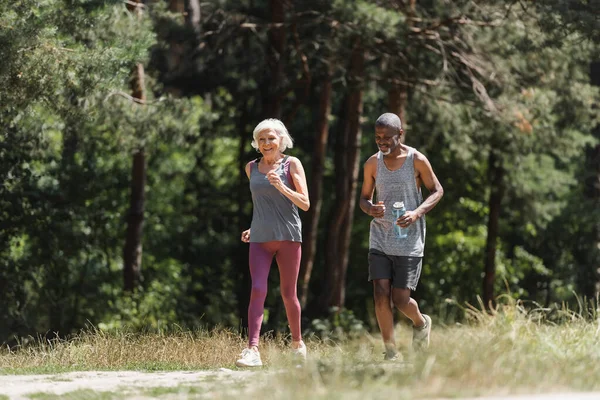 Senior sportswoman running near african american husband with sports bottle in forest