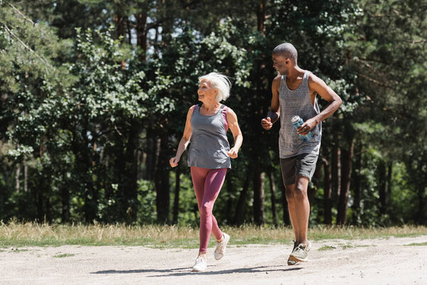 Smiling senior sportswoman jogging near african american husband with sports bottle in forest 