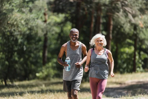笑着年长的女运动员跑到非洲裔美国丈夫身边 在森林里拿着运动瓶 — 图库照片