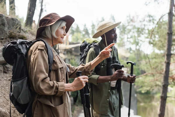 Senior tourist with trekking poles pointing with finger near blurred african american husband in forest