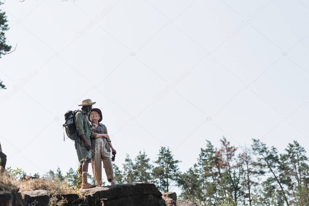 Senior woman smiling at african american husband with backpack on rock in forest 