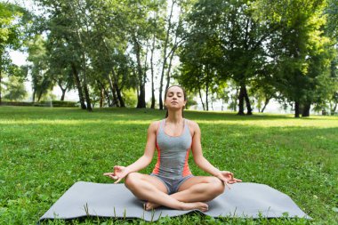 Gözleri kapalı genç bir kadın Lotus pozisyonunda oturuyor ve parkta yoga minderinde meditasyon yapıyor. 