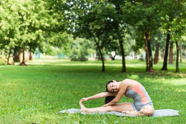 Spor giyimli esnek ve memnun bir kadın parkta yoga minderinde esniyor.