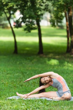 Spor giyimli esnek ve formda bir kadın parkta yoga minderinde esniyor.