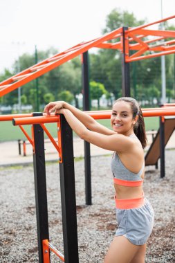 cheerful sportswoman smiling while leaning on parallel bars outside  clipart