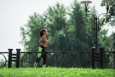 Parkta koşarken müzik dinleyen, kablosuz kulaklıklı mutlu ve sportif kadının yan görüntüsü