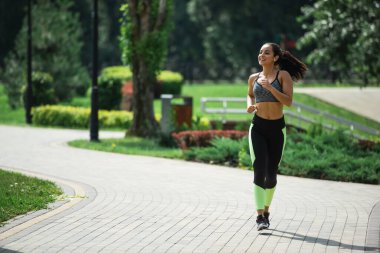 Kablosuz kulaklıkla parkta koşarken müzik dinleyen uzun boylu, güler yüzlü bir sporcu kadın.