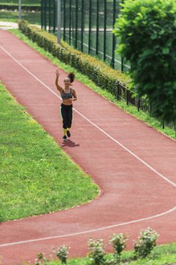 Kablosuz kulaklıklarla stadyumda koşarken el sallayan mutlu sporcu kadın.
