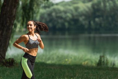 happy sportswoman in wireless earphones listening music while running in green park near lake clipart