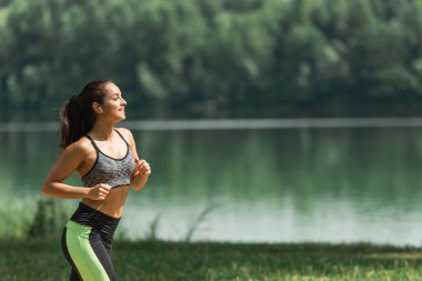 Göl kenarındaki yeşil parkta koşarken kulaklıkla müzik dinleyen mutlu sporcu kadının yan görüntüsü