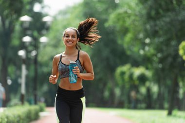 Yeşil parkta koşarken elinde spor şişesiyle kulaklık takan mutlu sporcu kadın.