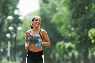 Kablosuz kulaklıklı neşeli sporcu kadın yeşil parkta koşarken elinde spor şişesi tutuyor.