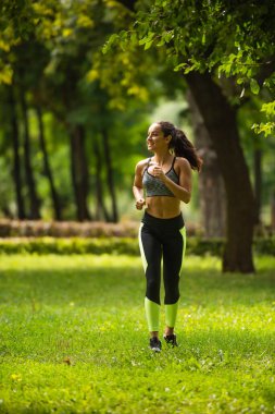full length of positive sportswoman in crop top and leggings jogging on grass in park  clipart