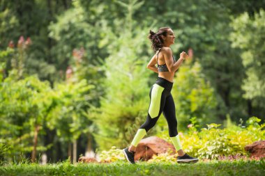 full length of joyful sportswoman in crop top and leggings jogging on grass in park  clipart