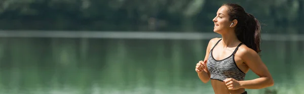 湖の近くの緑の公園で走りながら音楽を聴いているワイヤレスイヤフォンの幸せなスポーツウーマン バナー — ストック写真