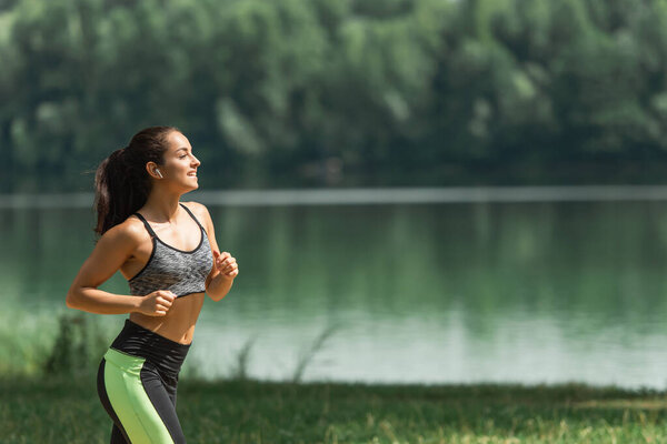 side view of happy sportswoman in wireless earphones listening music while running in green park near lake