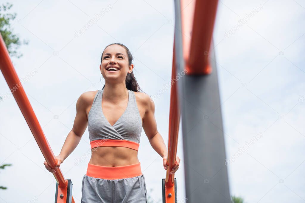 low angle view of cheerful and fit woman in sportswear exercising on parallel bars outside 