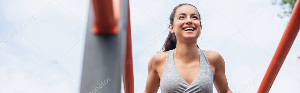 low angle view of cheerful and fit woman in sportswear exercising on parallel bars outside, banner