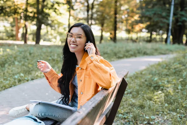 在公园的长椅上 拿着笔和笔记本在手机上聊天的快乐的亚洲学生 — 图库照片