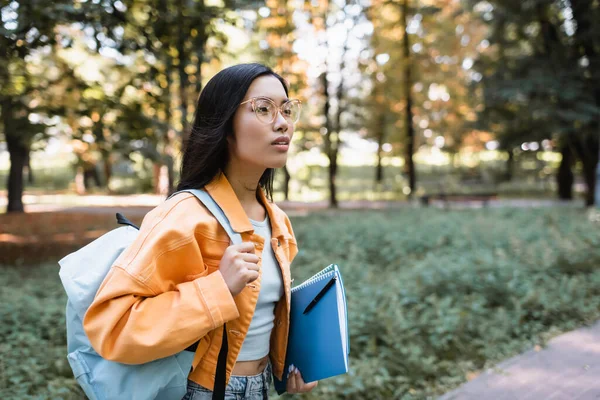 年轻的亚洲女人戴着眼镜 背着背包和笔记本在公园里走着 — 图库照片