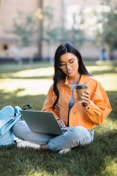 坐在草坪上边喝咖啡边用笔记本电脑笑着的亚洲女人 — 图库照片