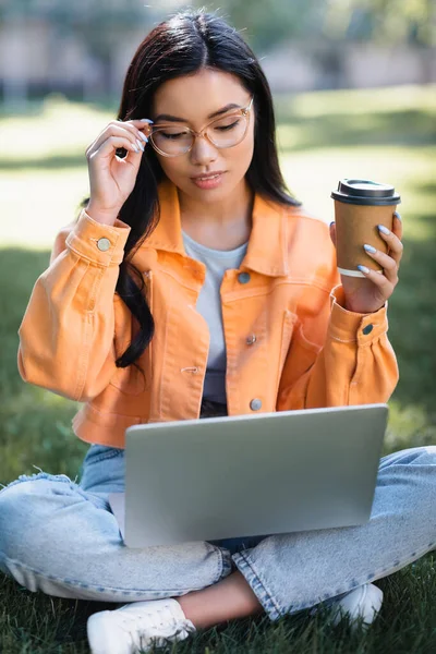 沉思的亚洲女人 一边看着笔记本电脑 一边拿着纸杯摸着眼镜 — 图库照片