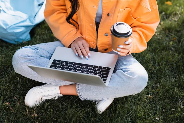 公園で芝生の上でノートパソコンを使ってオレンジのジャケットやジーンズを着た生徒の — ストック写真