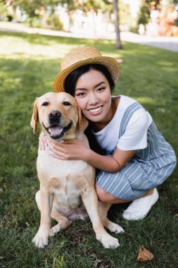 Genç Asyalı kadın parkta köpeğe sarılırken kameraya gülümsüyor.